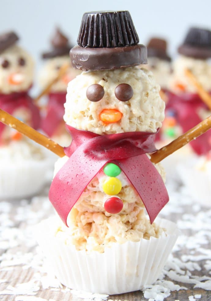 close up picture of a snowmen rice krispy treat with a peanut butter cup hat and tiny m&ms for buttons and eyes. One snowman in the front with four snowmen in the background.