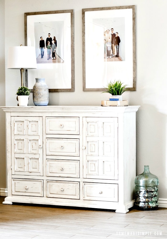 a large white cabinet with a lamp, pot and small plant on top. Above the cabinet are two family pictures.