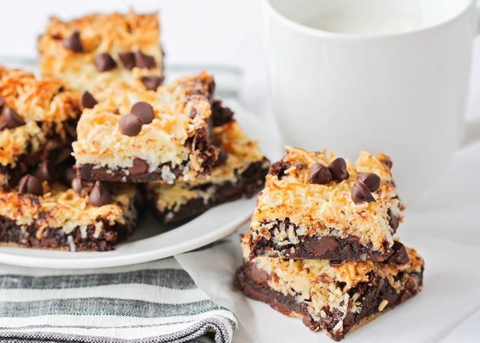 a white plate full of Toasted Coconut Dark Chocolate Brownies. Next to the plate are two more squares of coconut dark chocolate brownies on a napkin with a white mug filled with milk behind them.