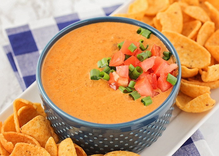 a blue bowl filled Chili Cream Cheese Dip topped with diced tomatoes and green onions on a serving tray with corn chips next to it