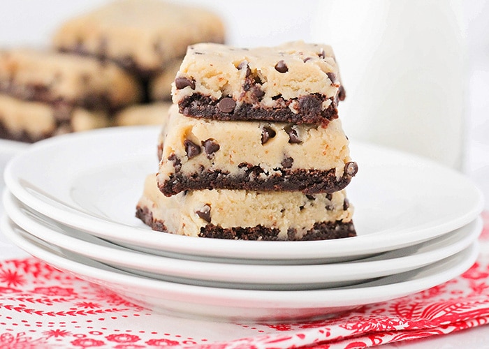 three Cookie Dough Brownies stacked on top of each other sitting on a stack of three white plates. The plates are on top of a red and white cloth napkin and a plate of more cookie dough brownies and a glass pitcher of milk are in the background.