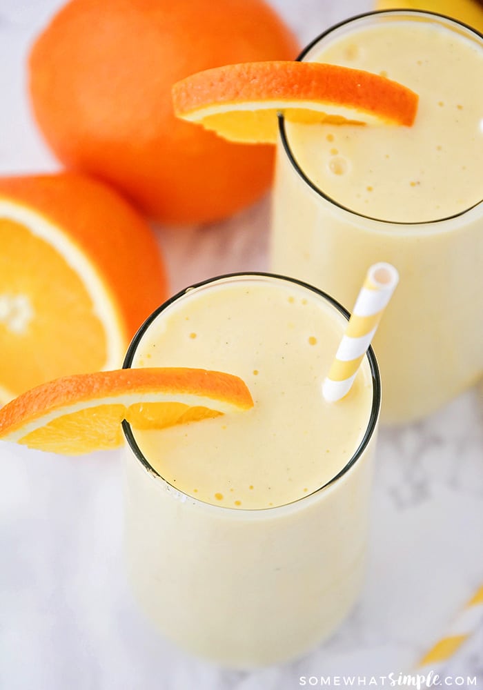 looking down on two orange creamsicle drinks with a slice of orange on the rim of each glass. A striped orange straw is in one of the glasses and two oranges are on the counter next to the drinks.