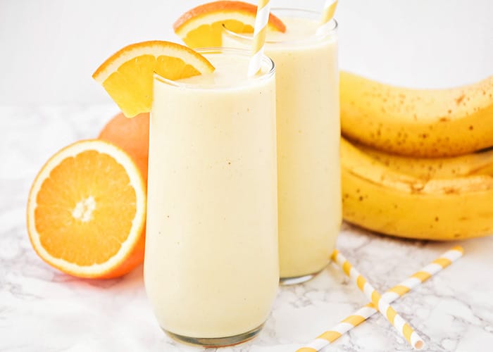 two glasses of this frozen orange creamsicle smoothie on a counter with oranges and bananas in the background and two striped orange straws on the counter next to the glasses