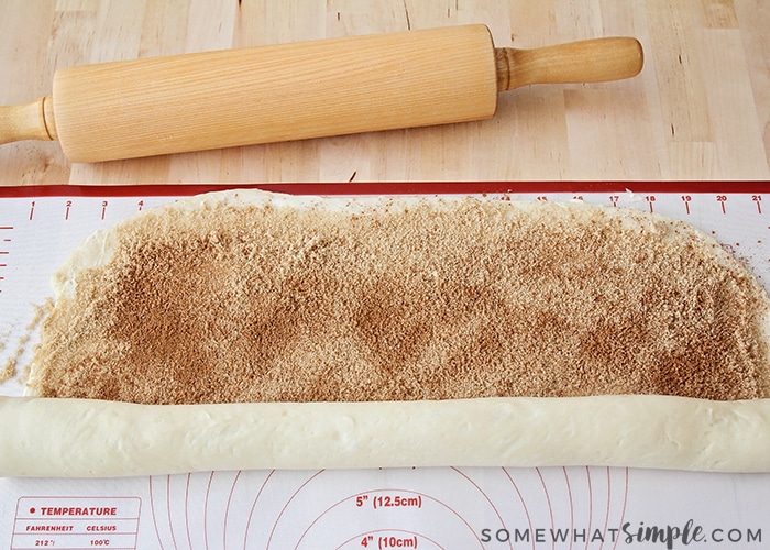 cinnamon roll dough that has been rolled out with cinnamon and sugar sprinkled over it and is beginning to be rolled up with a rolling pin at the top of the image