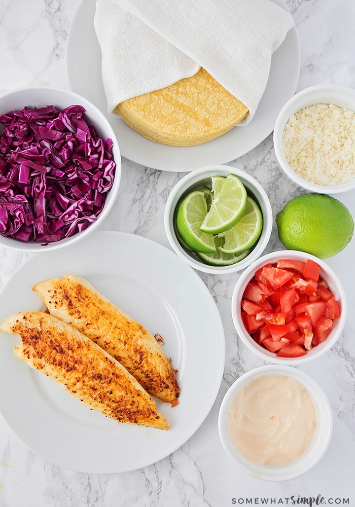 Looking down on the ingredients to make fish tacos; baked tilapia, purple cabbage, corn tortillas, lime slices, diced tomatoes, fish taco sauce and cheese