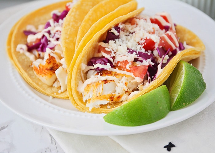 two tilapia fish tacos in corn tortillas topping with lettuce tomatoes and cheese on a white plate with two slices of lime on the plate next to the tacos.