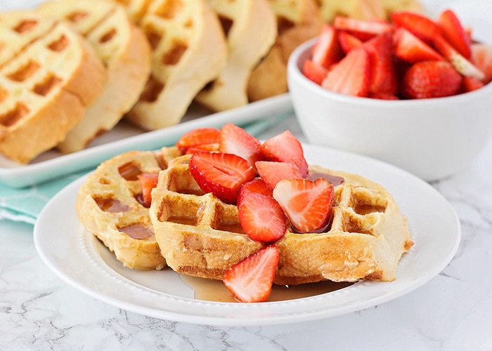 a white plate with two French toast waffles on it that are topped with maple syrup and sliced strawberries. A bowl of sliced strawberries and a tray of more french toast waffles are in the background.