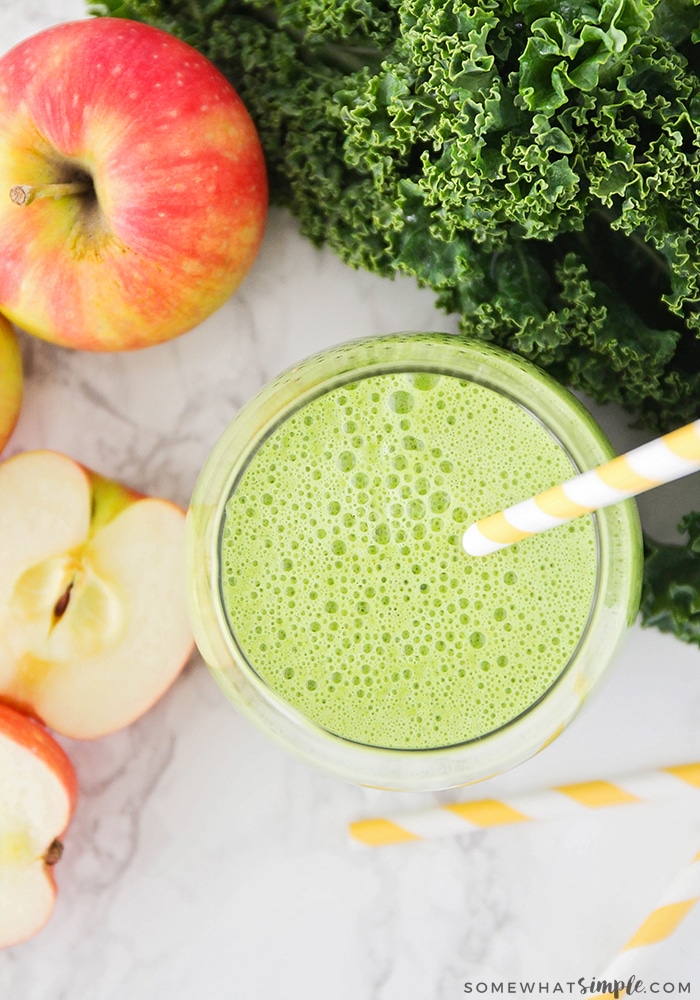 looking down into a glass filled with this simple green smoothie recipe and a yellow striped straw in the cup. Next to the glass on the counter are apples, leaves of kale and an additional striped yellow straws.