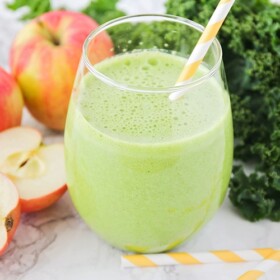 a glass filled with a green smoothie and a yellow striped straw in the cup. Next to the glass on the counter are apples, leaves of kale and 2 more striped yellow straws.