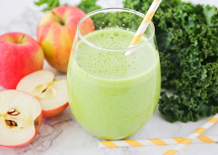 a glass filled with a green smoothie and a yellow striped straw in the cup. Next to the glass on the counter are apples, leaves of kale and 2 more striped yellow straws.