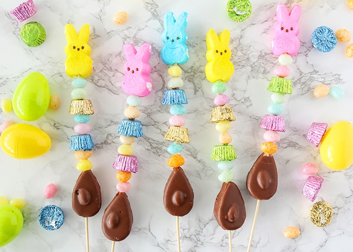 five Easter candy kababs laying on a white counter that are made with chocolate eggs, peanut butter cups, jelly beans and marshmallow candy. Plastic eggs, and additional candy is scattered around them on the counter.