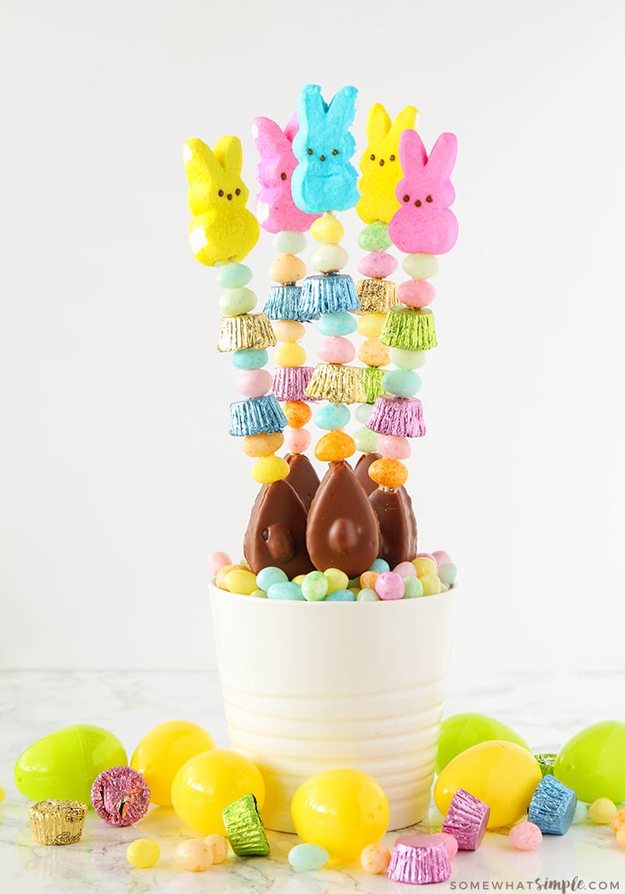 five candy kabobs are standing up in a white bowl that is filled with pastel colored candies. The kabobs are made with various pieces of Easter candy. Plastic eggs are on the counter around the bowl.