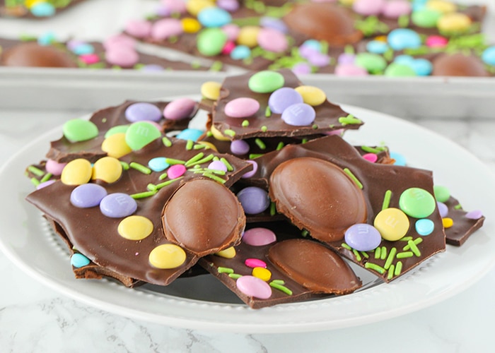 a white plate filled with pieces of dark chocolate bark topped with pastel colored candy, sprinkles chocolate eggs. Behind the plate is a cookie sheet filled with more pieces of Easter bark.