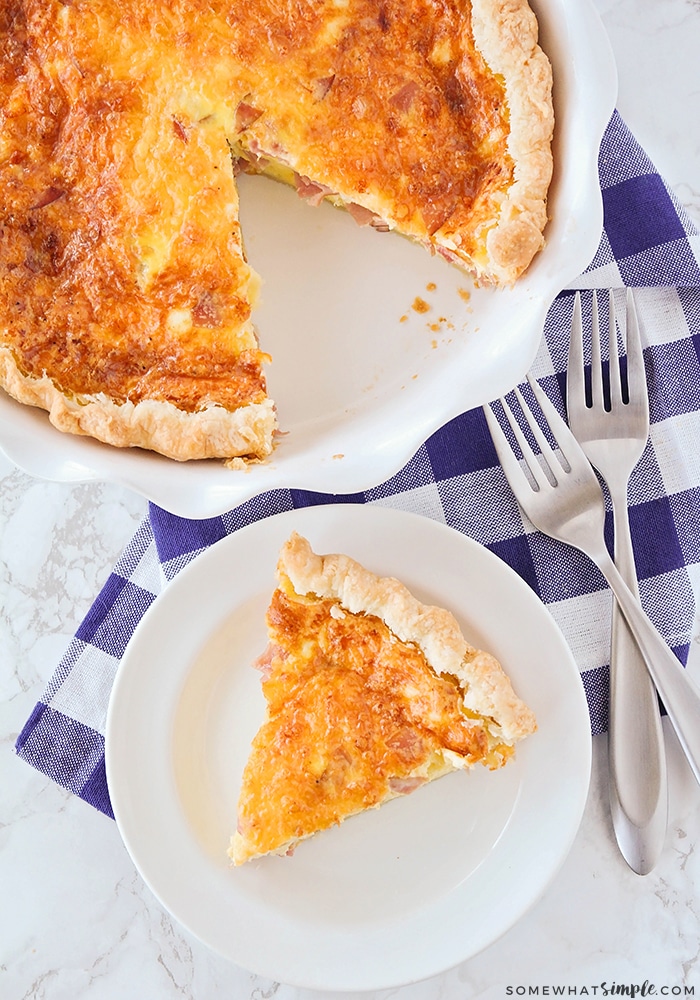 A full breakfast quiche in a white baking dish with a slice cut out. Next to the baking dish is a slice of ham and cheese quiche.