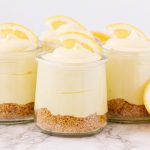 four cups of lemon cheesecake mousse topped with lemon slices and lemons next to the cups on the counter