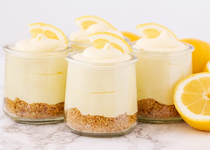 four cups of lemon cheesecake mousse topped with lemon slices and lemons next to the cups on the counter