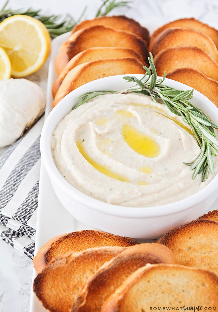 a close up of a serving tray with slices of toasted bread. In the middle of the tray is a white bowl filled with hummus.