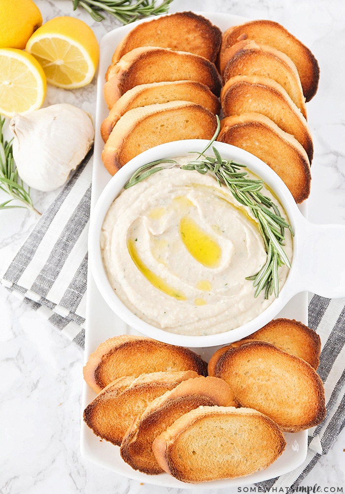 looking down on a serving tray filled with slices of toasted baguette. In the center of the tray is a white bowl filled with this easy homemade hummus recipe. A branch of rosemary has been laid on top of the hummus. Next to the serving tray are 3 lemons and a head of garlic.