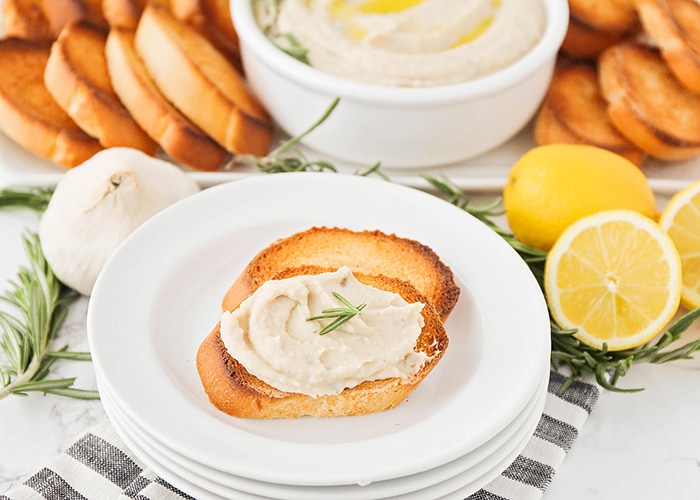 two pieces of toasted baguettes on a stack of white plates topped with Homemade Hummus. Behind the plates is a serving tray with more slices of toasted slices of bread with a white bowl of hummus. There are lemons, an onion and leaves of rosemary on the counter next to the plates.