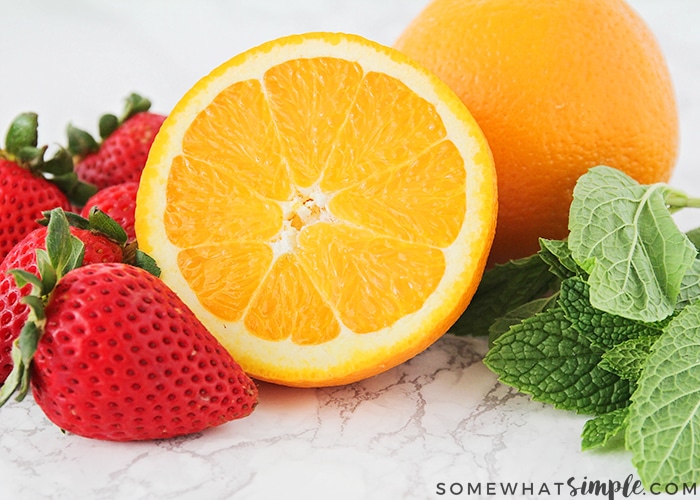 whole bright red strawberries an orange sliced in half with a full orange behind it and mint leaves sitting on a white counter. These are the ingredients for this recipe.