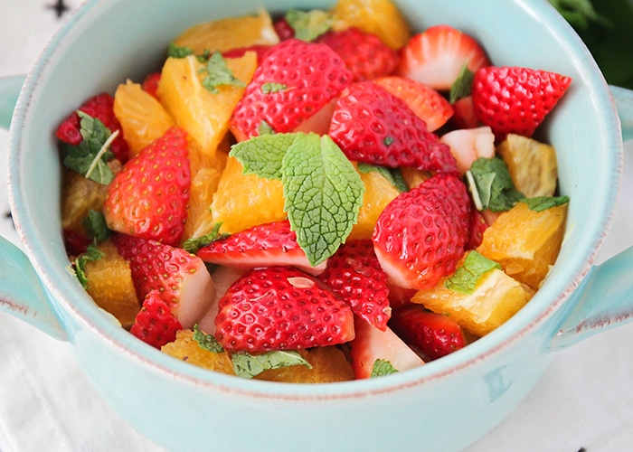 a light blue bowl filled with strawberry mint fruit salad and oranges topped with small mint leaves.