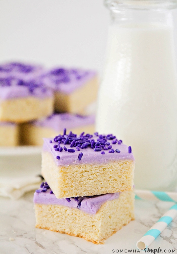 two sugar cookie bars are stacked on each other sitting on the counter top. The cookie bars are topped with purple frosting and purple sprinkles. On the counter behind the bars is a pitcher of milk and a plate filled with sugar cookie bars. 