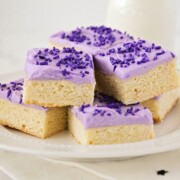a white plate full of sugar cookie bars that are stacked on top of each other. Each cookie bar is topped with purple frosting and purple sprinkles. A glass of milk is on the counter behind the plate of sugar cookie bars.