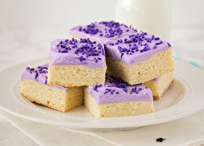 a white plate full of sugar cookie bars that are stacked on top of each other. Each cookie bar is topped with purple frosting and purple sprinkles. A glass of milk is on the counter behind the plate of sugar cookie bars.