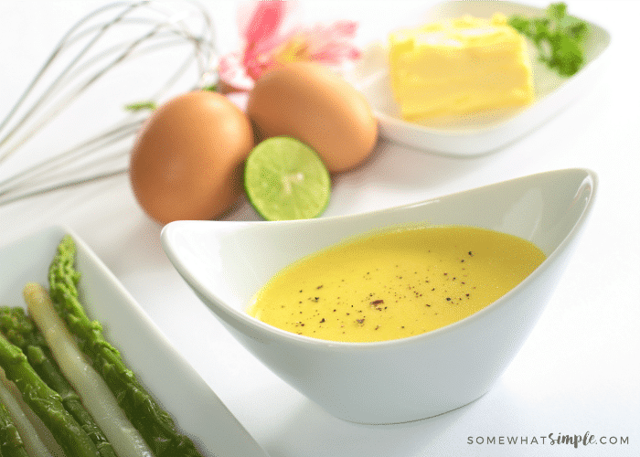 a white dish of Hollandaise Sauce made in a blender with this easy Recipe with a plate of asparagus next to it and eggs and butter in the background