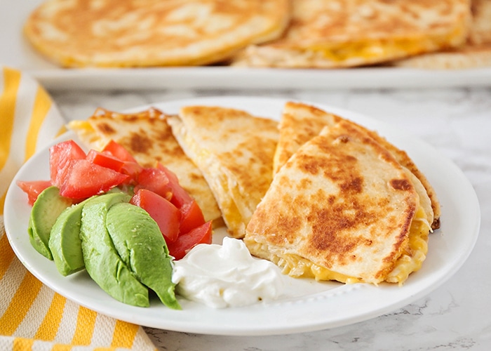 Cheesy Chicken Quesadillas on a plate with a side of sour cream, avocado and tomatoes are a popular super bowl snack