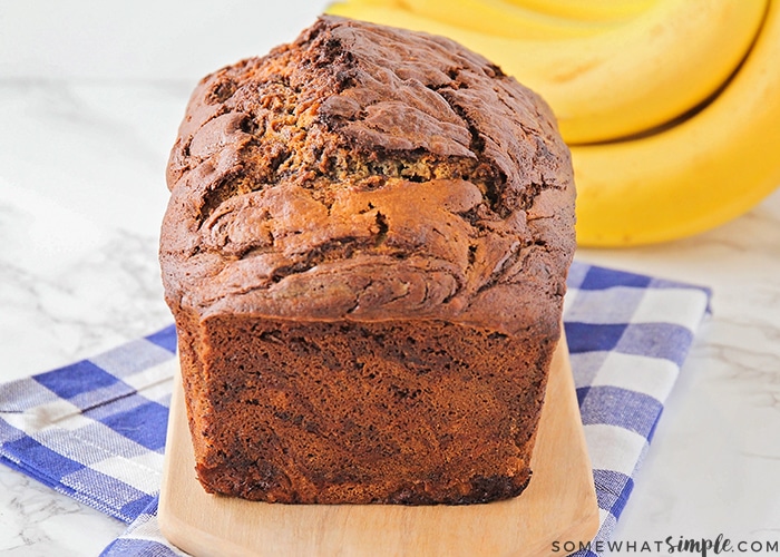 a baked loaf of chocolate banana bread on a counter made using this easy recipe with a bunch of bananas on the counter in the background