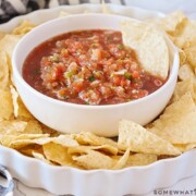 a white bowl filled with this easy blender salsa recipe with a tortilla chip standing inside the bowl. This bowl is in the center of a white serving dish with tortilla chips all around the bowl.