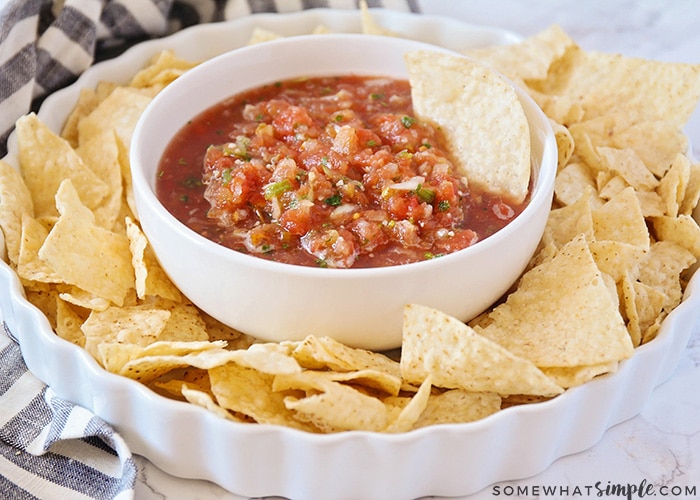 a white bowl filled with this easy blender salsa recipe with a tortilla chip standing inside the bowl. This bowl is in the center of a white serving dish with tortilla chips all around the bowl.