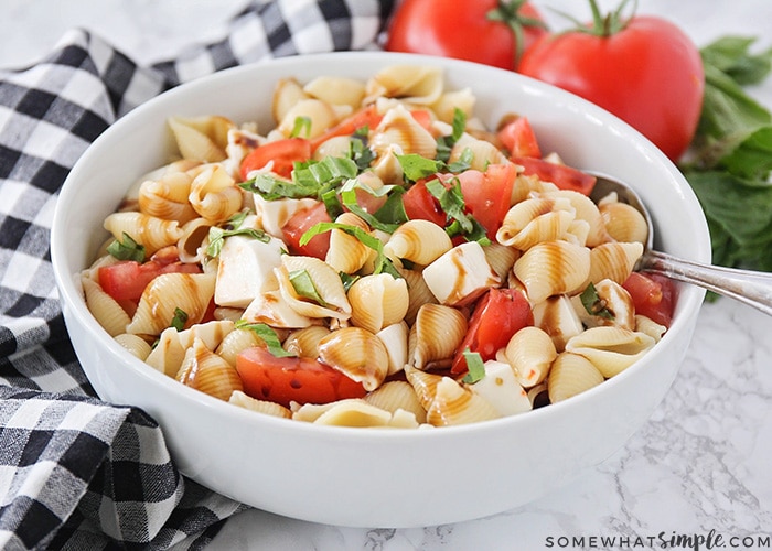 a white bowl filled with a caprese pasta salad that is made with shell pasta, fresh mozzarella, tomatoes and basil, tossed in italian dressing and topped with a balsamic glaze. A spoon has been set in the bowl and two tomatoes and fresh basil are in the background and a black and white checkered cloth napkin is on the counter next to the bowl.