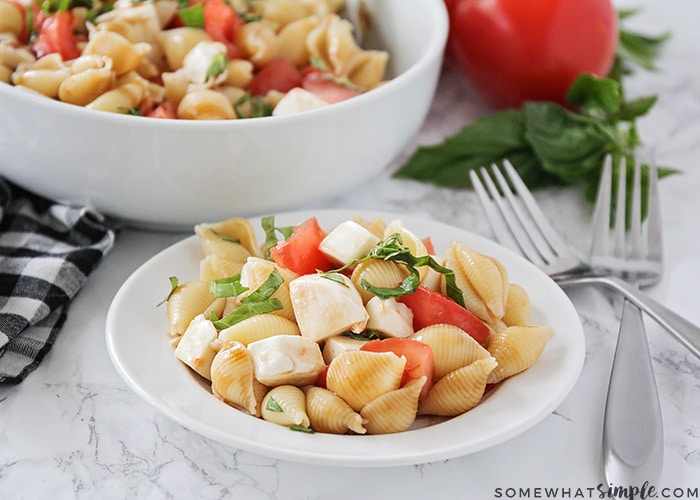 a white plate filled with this caprese pasta salad recipe. The ingredients used to make it were shell pasta, fresh mozzarella, tomatoes and basil, tossed in italian dressing and topped with a balsamic glaze. Two forks on on the counter next to the plate and a bowl of more caprese salad is in the background with a red tomato next to it.
