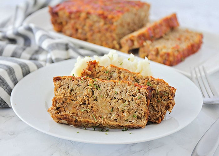 two slices of meatloaf on a white plate made with this simple meatloaf recipe. A loaf on a serving tray is in the background.