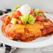 two enchilada style baked bean burritos on a white plate topped with lettuce, tomatoes and sour cream. Next to the plate is a black and white checkered cloth napkin.