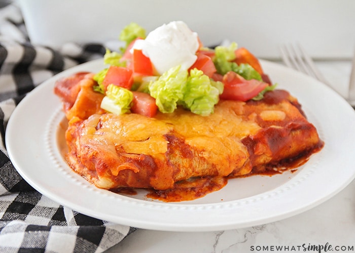 two enchilada style baked bean burritos on a white plate topped with lettuce, tomatoes and sour cream. Next to the plate is a black and white checkered cloth napkin.