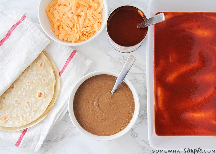 a white tray with enchilada sauce lining the bottom, a bowl of shredded cheese, a dish of refried beans, a can of red enchilada sauce and a stack of flour tortillas are the ingredients you'll need to make these baked bean burritos