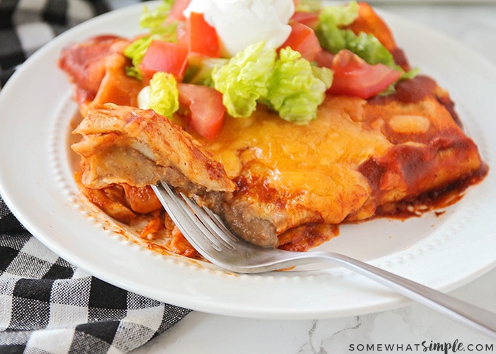 two enchilada style baked bean burritos on a plate topped with lettuce and tomatoes. A fork has cut a bit off the corner and is laying on the plate with the piece of burrito stuck to the fork.