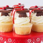 a red cake stand with seven graduation cupcakes on it. They are vanilla cupcakes with vanilla frosting that have graduation toppers made with peanut butter cups, squares of chocolate, red candies and thin red licorice