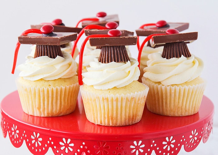 a red cake stand with seven graduation cupcakes on it. They are vanilla cupcakes with vanilla frosting that have graduation toppers made with peanut butter cups, squares of chocolate, red candies and thin red licorice