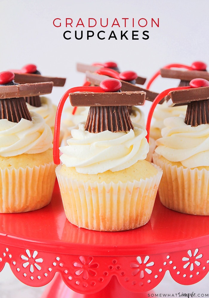 a close up of these graduation cupcakes on a red cake stand. They are vanilla cupcakes with vanilla frosting that have graduation toppers made with peanut butter cups, squares of chocolate, red candies and thin red licorice with the words graduation cupcakes written at the top of the image