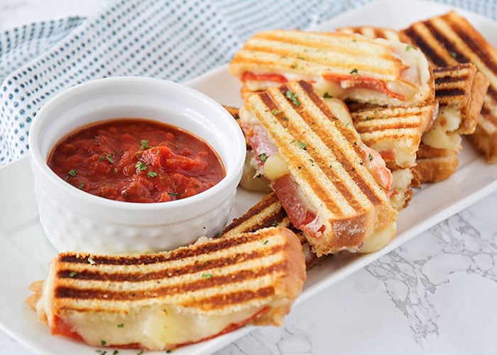 a long white serving tray filled with several grilled cheese pizza dippers filled with mozzarella cheese and pepperonis and a small white bowl filled with red marinara sauce on the tray 