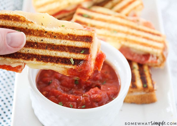 a hand holding a slice of a grilled pizza dipper that has just been dipped in marinara sauce. Below the cheese dipper is a white tray with a small bowl of marinara sauce and additional slices of pizza dippers