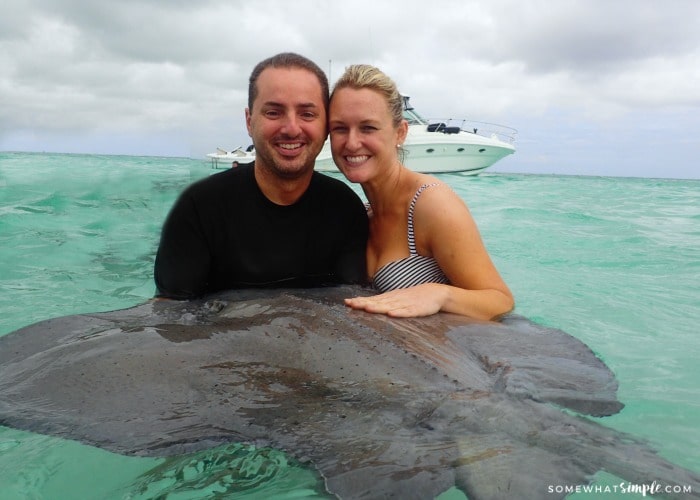 a beautiful blonde woman in a bikini with her husband in the ocean holding a large sting ray with a boat in the background.