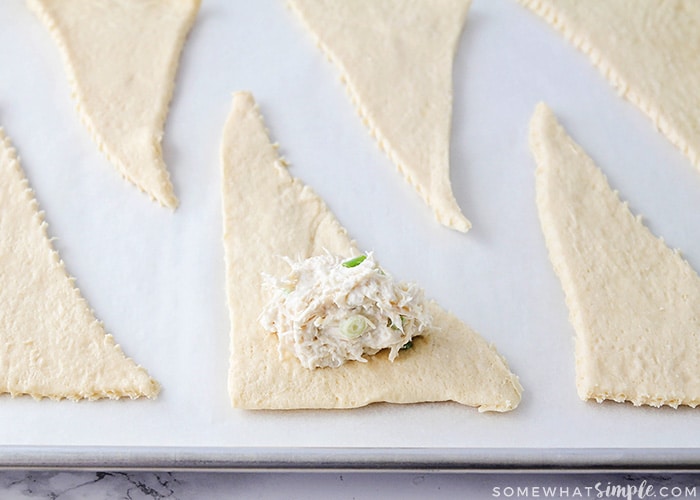 triangles of crescent roll dough layed out on a baking sheet. One of the crescent rolls has a scoop of the chicken and cheese mixture that goes inside these roll ups.