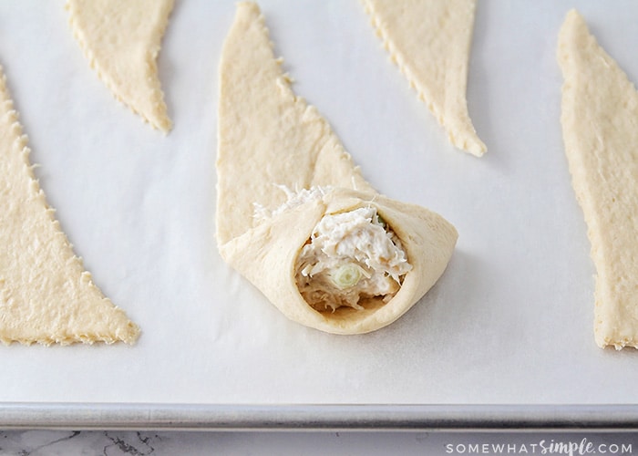 triangles of crescent dough layed out on a baking sheet. the one in the middle has a scoop of chicken mixture and the two corners of the dough has been folded over the top of the mixture.