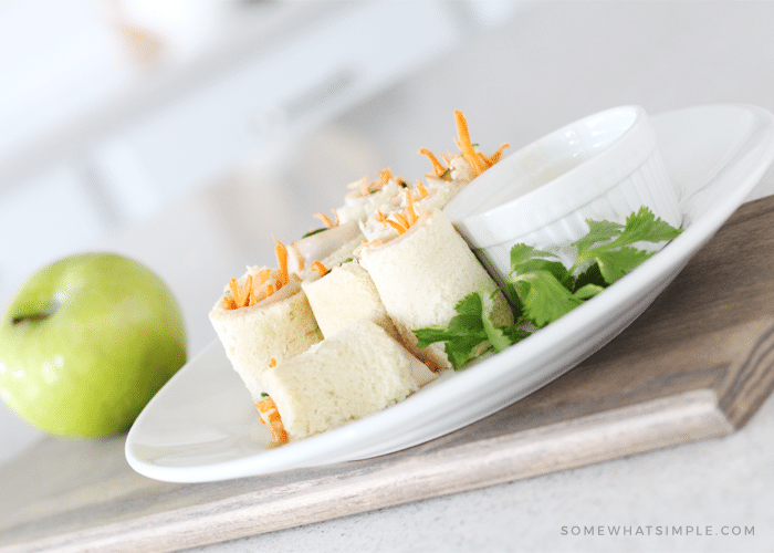 a white plate with several sandwich sushi rolls with a side of ranch sitting on a cutting board with a green apple next to the plate