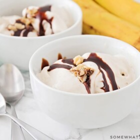 two white bowls filled with frozen banana ice cream topped with chocolate syrup and nuts. On the counter next to the bowls are two spoons and a small bunch of bananas